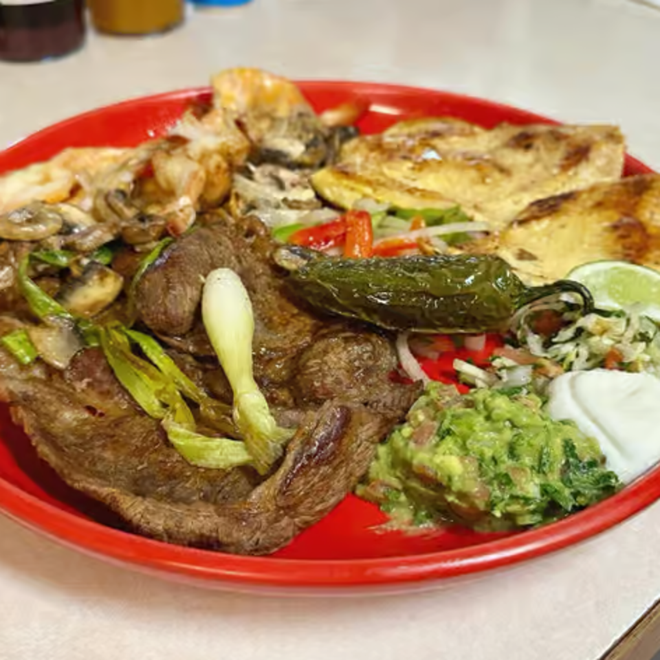 Mexican platter with grilled steak, vegetables, roasted pepper, tortillas, guacamole, sour cream, and lime on a red plate.