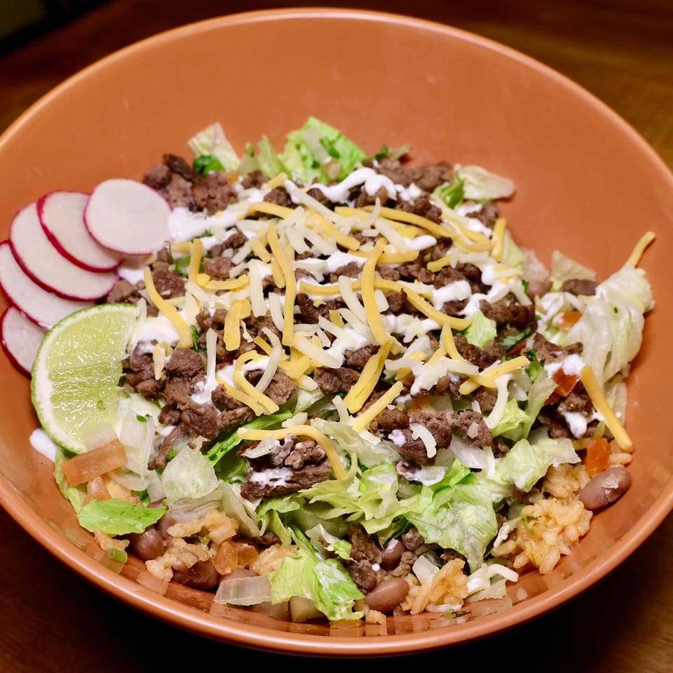 Taco salad with ground beef, shredded lettuce, cheese, sour cream, radishes, and lime in a brown bowl.