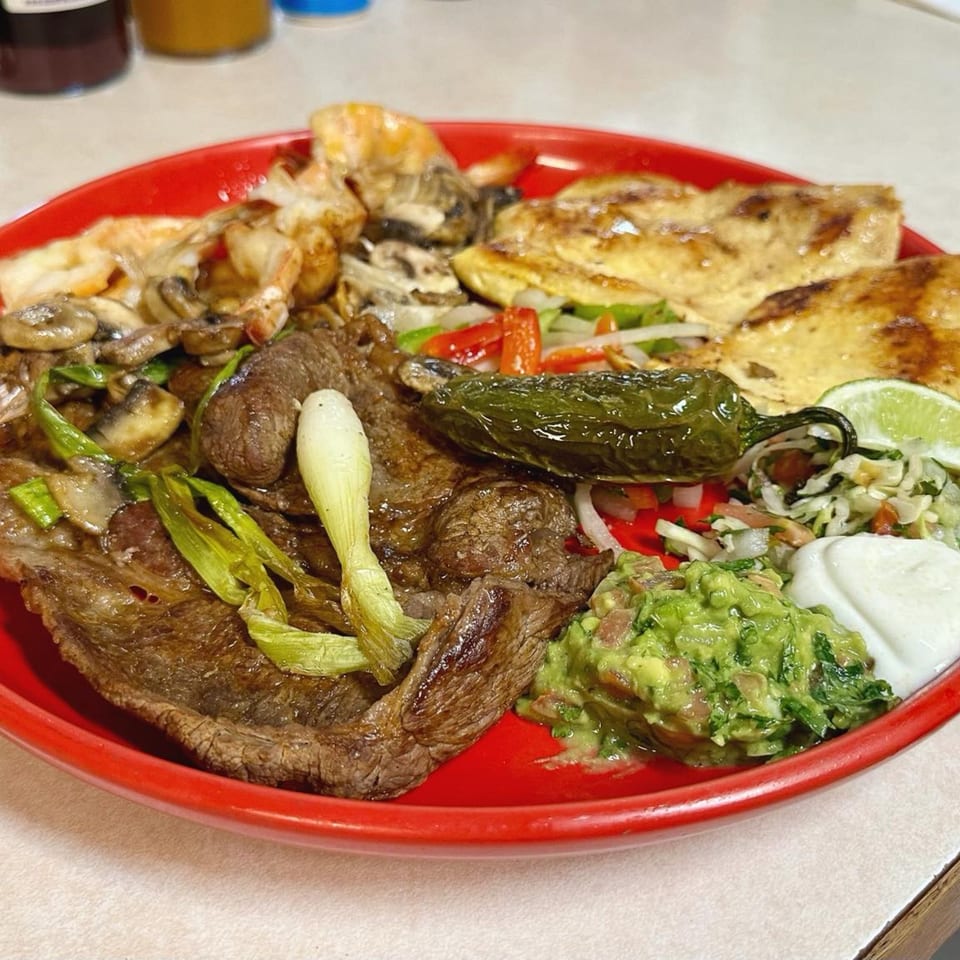 A vibrant red plate filled with grilled steak, chicken, mushrooms, guacamole, sour cream, and a jalapeño.